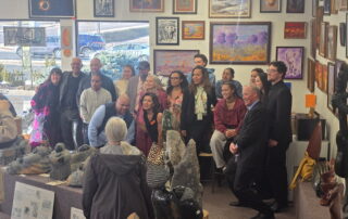 Rex Mhiripiri's family poses together during a Mhiripiri Gallery event, surrounded by Shona stone sculptures and colorful framed artwork. A large collection of carved stone figures fills the foreground while the group gathers at the back of the gallery.