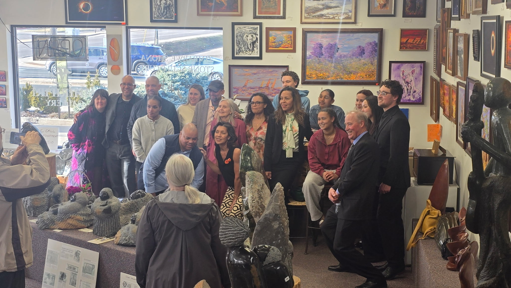 Rex Mhiripiri's family poses together during a Mhiripiri Gallery event, surrounded by Shona stone sculptures and colorful framed artwork. A large collection of carved stone figures fills the foreground while the group gathers at the back of the gallery.