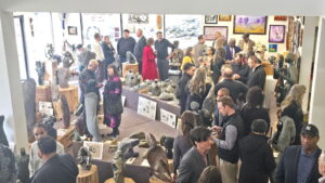 A busy Mhiripiri Gallery event with guests gathering and talking among displays of Zimbabwean Shona stone sculptures and colorful paintings. Sculptures fill tables and wooden pedestals throughout the gallery.